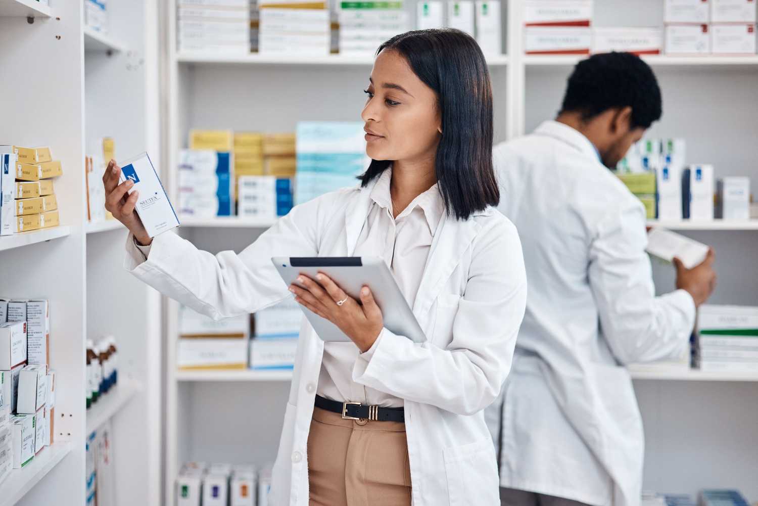 A woman pharmacist holding a clipboard and looking at a box of medicine from the stock shelves